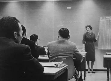 An old black and white image of SSE founder, Susan S. Elliott at the front of a classroom by a chalkboard, instructing a room of tech professionals.
