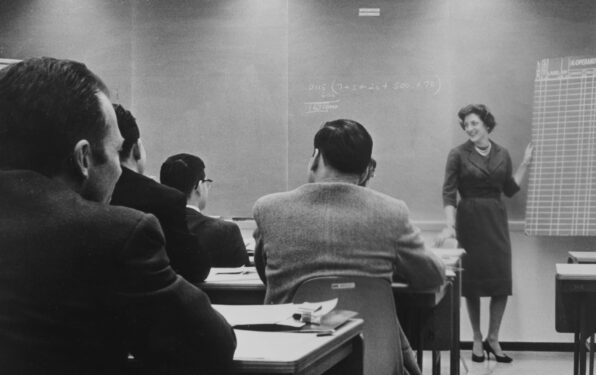 An old black and white image of SSE founder, Susan S. Elliott at the front of a classroom by a chalkboard, instructing a room of tech professionals.