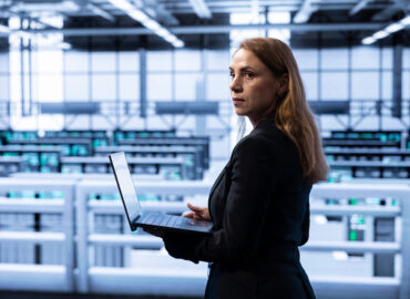 A woman standing on an elevated platform in a server room, holding a laptop while peering out toward the left of the camera.