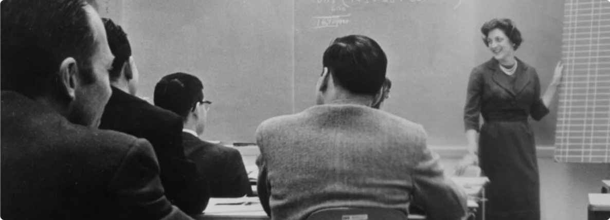 An old, black and white photo of SSE founder, Susan S. Elliott, standing at the front of a class in front of a chalkboard, instructing tech professionals.