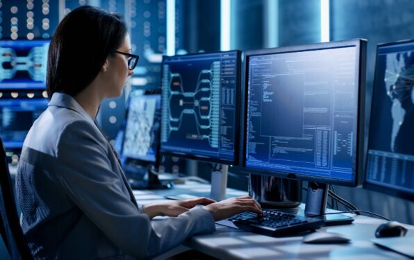 Female IT professional sitting at a desk with three monitors, left showing connection map, center with code, and right showing global map with various connection points.