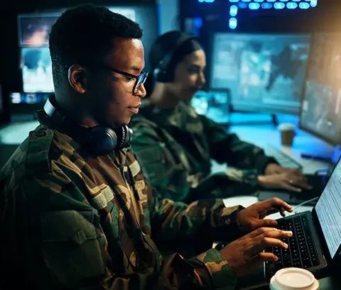 An armed services veteran working at a laptop on an aerospace training system, with another veteran seated in the background working on a separate laptop near several large monitors.