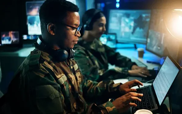 An armed services veteran working at a laptop on an aerospace training system, with another veteran seated in the background working on a separate laptop near several large monitors.