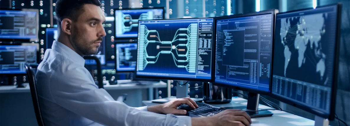A male CMMC expert seated at a desk, analyzing the data across three large monitors: one with a connections chart, another with code, and another with a world map.