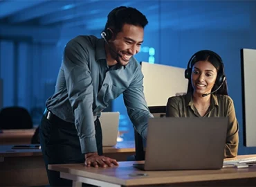 Two IT professionals at a desk, the male standing and leaning toward a laptop, the woman seated and looking at the laptop, both smiling and wearing headsets.