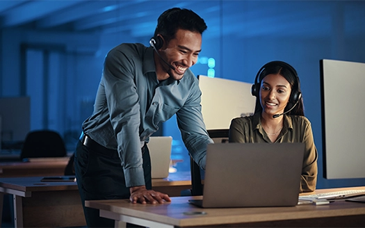 Two IT professionals at a desk, the male standing and leaning toward a laptop, the woman seated and looking at the laptop, both smiling and wearing headsets.