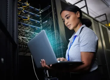 A female IT professional looks at data on a laptop that is plugged directly into a server cabinet to her right.