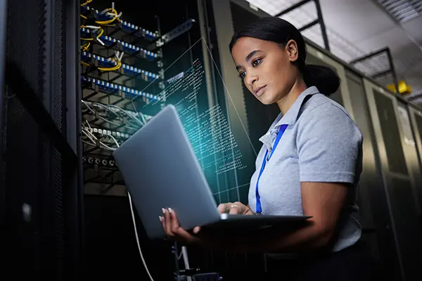 A female IT professional looks at data on a laptop that is plugged directly into a server cabinet to her right.