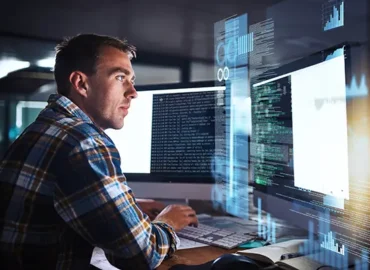 Male CMMC specialist working at his desk with multiple monitors, looking at one with various code on the screen as well as stylized graphs and data imagery surrounding the screen.