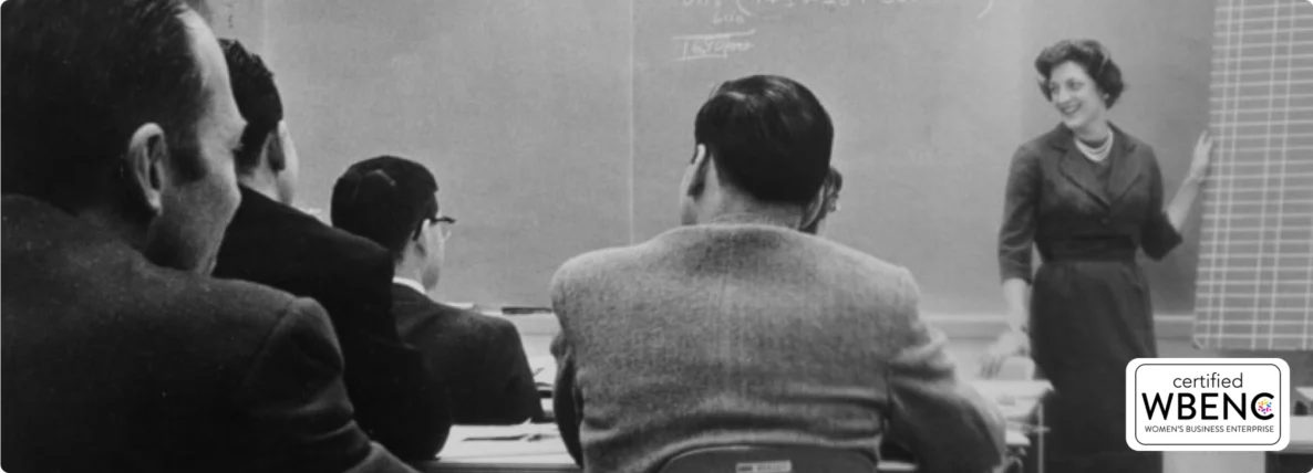 Old black and white photo of SSE founder, Susan S. Elliott, standing at a chalkboard in front of a classroom of tech professionals, with the Certified WBENC Women's Business Enterprise badge in the bottom right corner.