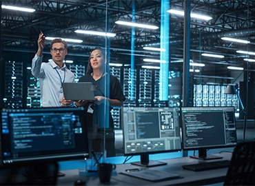 A male and female professional standing inside the glassed-in area of servers, holding a laptop, pointing and looking in toward a work area with monitors displaying data.