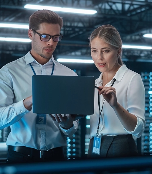 Man and woman wearing professional attire and badges, working together in a server room, looking at a laptop to verify security.
