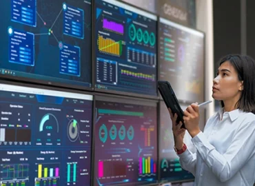 A young woman in professional attire with a tablet and stylus taking notes from the various screens of data displayed in front of her on a wall.