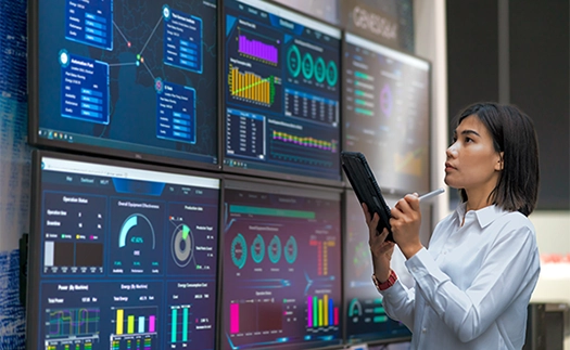 A young woman in professional attire with a tablet and stylus taking notes from the various screens of data displayed in front of her on a wall.